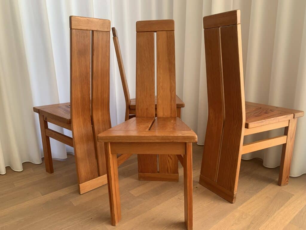 Vier houten eetkamerstoelen met hoge, rechthoekige rugleuningen staan in een halve cirkel op een lichte houten vloer, voor witte, tot op de vloer reikende gordijnen.