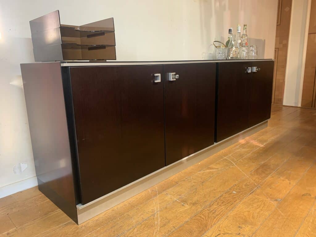 Een donkerhouten dressoir met zilveren handgrepen staat op een houten vloer. Links staan er een stapel dienbladen en rechts een assortiment flessen en glazen. De muur erachter is effen en lichtgekleurd.