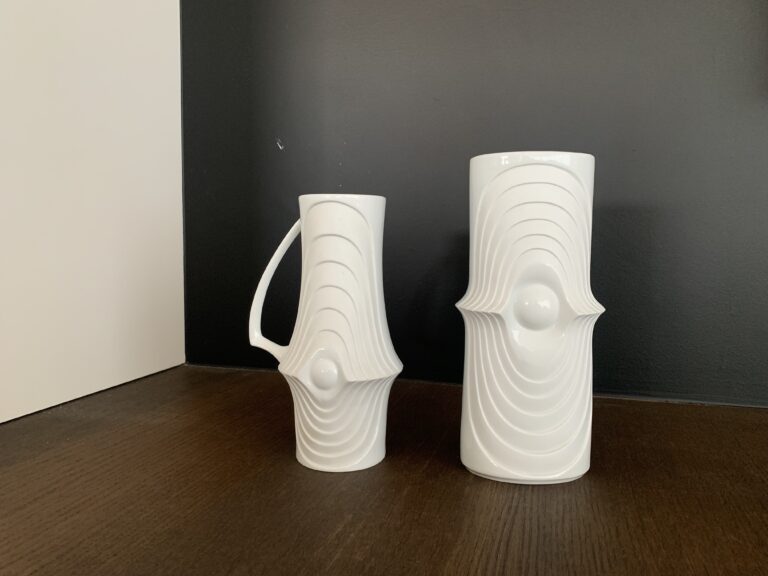 Twee witte keramische vazen met geometrische, concentrische reliëfpatronen en centrale uitstulpingen staan op een donker houten oppervlak tegen een zwart-witte muurachtergrond.