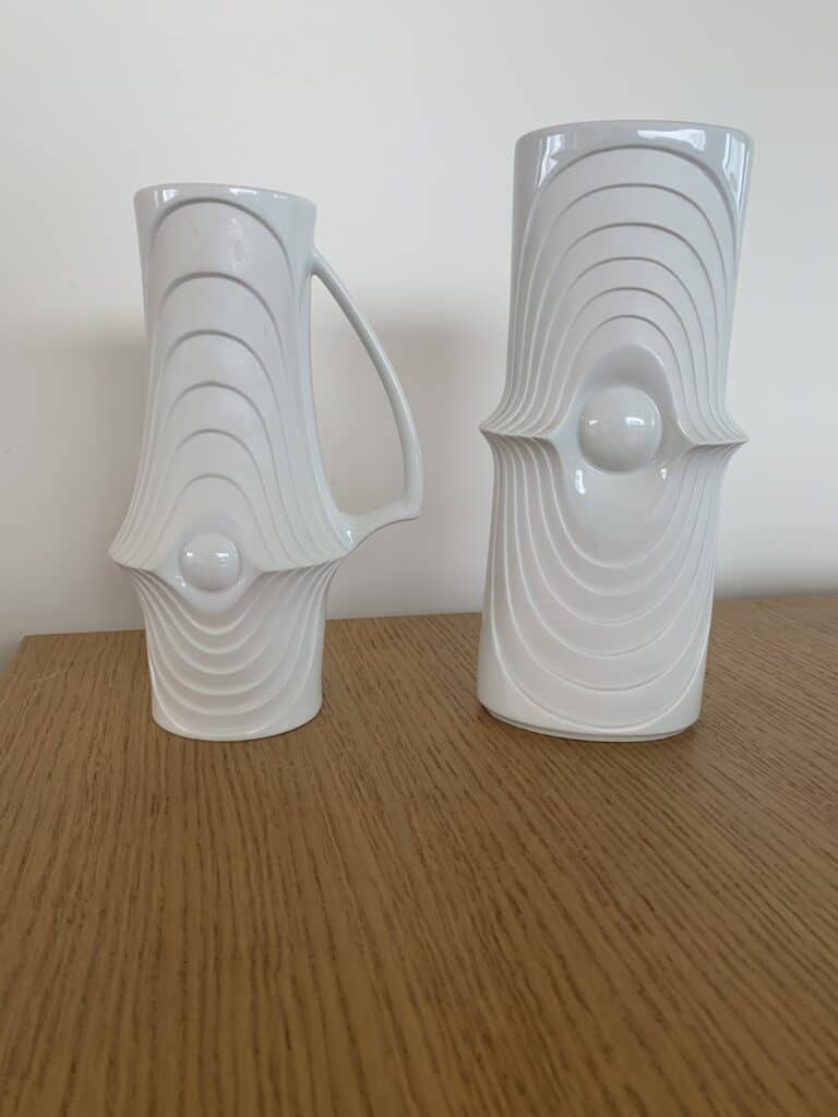 Twee witte keramische vazen met abstracte, golvende, verhoogde patronen en centrale ronde uitstulpingen, geplaatst op een houten oppervlak tegen een effen lichtgekleurde muur.