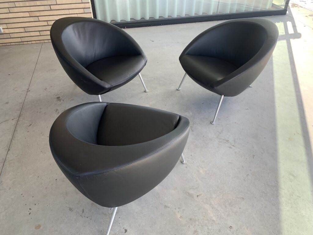 Drie moderne, zwarte, ronde stoelen met metalen poten staan in een losse driehoek op een grijze betonnen vloer, naast een bakstenen muur en een groot raam met een gordijn.