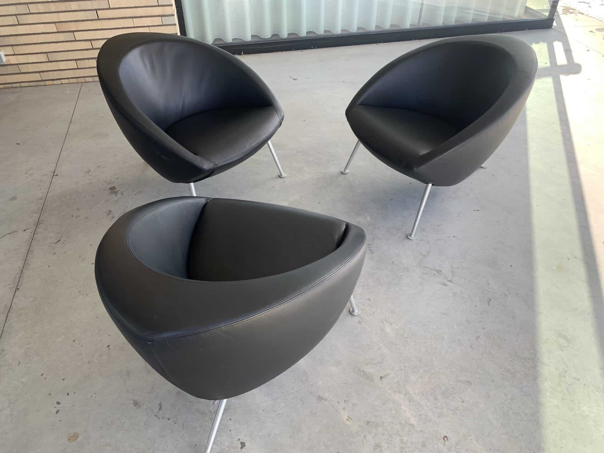 Drie moderne, zwarte, ronde stoelen met metalen poten staan in een losse driehoek op een grijze betonnen vloer, naast een bakstenen muur en een groot raam met een gordijn.