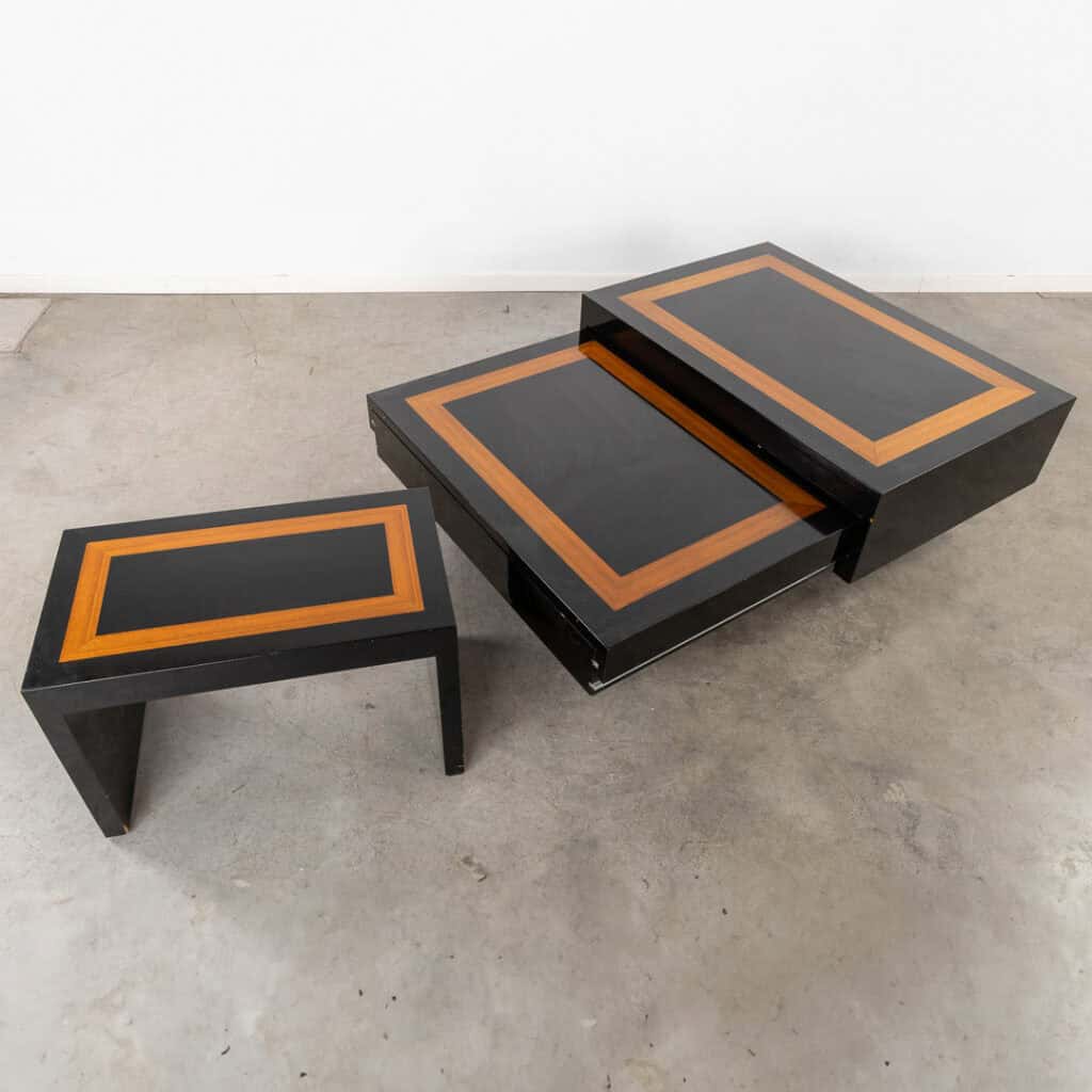 Een set moderne, zwarte bijzettafeltjes met rechthoekige oranje houten inlegstukken, tentoongesteld op een betonnen vloer. Eén tafel is gedeeltelijk uitgeschoven en de andere is volledig losgemaakt.