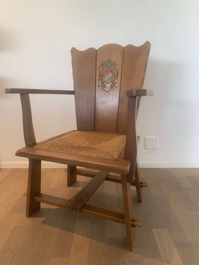 Een houten fauteuil met een hoge rugleuning met een gebeeldhouwd en beschilderd wapen, gevlochten zitting en geometrische poten, geplaatst op een lichte houten vloer tegen een witte muur.