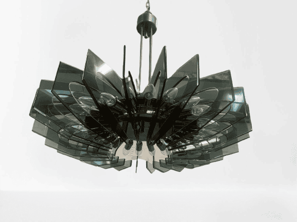 Een moderne kroonluchter met gelaagde, rechthoekige, donkere glaspanelen in een cirkelvormig patroon rond een centraal messing armatuur, hangend aan het plafond.