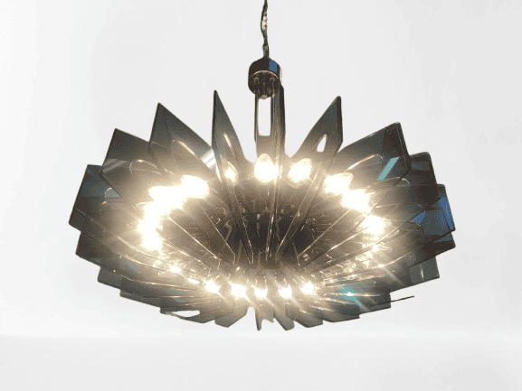 Een moderne kroonluchter met meerdere driehoekige glaspanelen die in een cirkelvormig patroon zijn gerangschikt en helder licht uitstralen via verschillende gloeilampen. De kroonluchter hangt aan het plafond tegen een effen witte achtergrond.