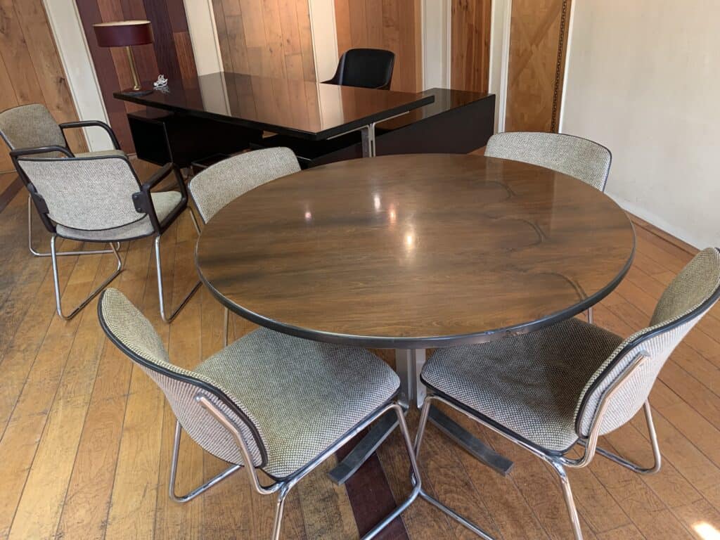 Een ronde houten tafel met vier grijze stoelen staat op een houten vloer. Op de achtergrond staat een groter donker bureau met een zwarte stoel en een lamp. De setting lijkt een kantoor of vergaderruimte te zijn.