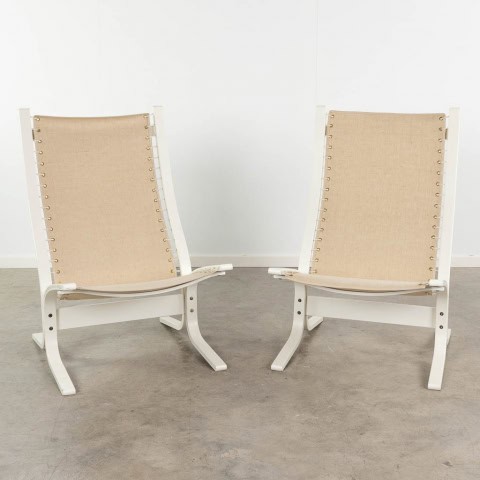 Twee moderne loungestoelen met een wit houten frame en beige canvas zittingen staan naast elkaar op een betonnen vloer tegen een eenvoudige witte muur.