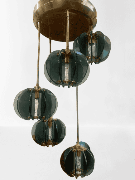 Een moderne kroonluchter met vijf zichtbare gloeilampen, elk omgeven door groene, bolvormige glaspanelen, hangt aan een ronde plafondbevestiging van messing.