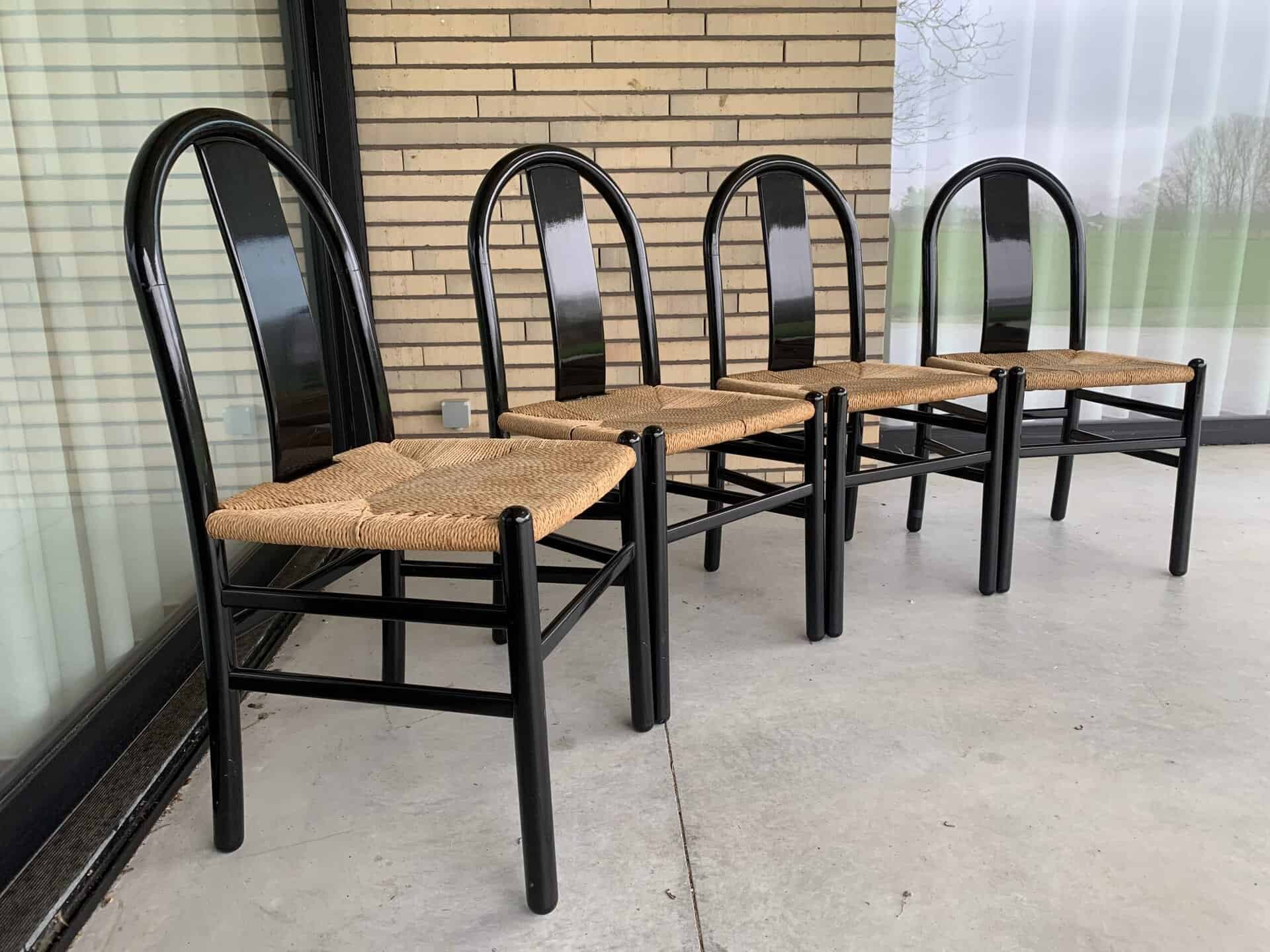 Vier zwarte metalen stoelen met gevlochten rieten zittingen staan op een rij op een grijs betonnen terras, voor een gebouw met lichtgekleurde bakstenen muren en grote glazen ramen.