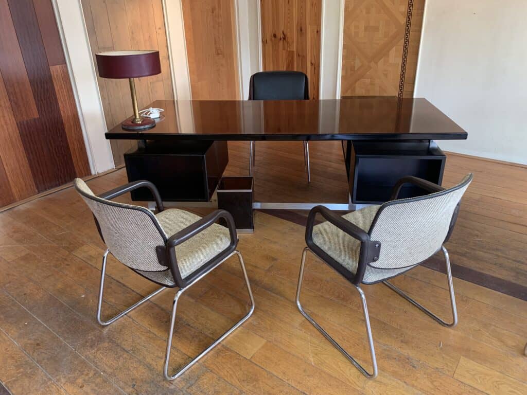 Een groot, donkerhouten bureau met een zwarte stoel erachter en twee beige stoelen met metalen frame ervoor. Op het bureau staan een bruine bureaulamp en een draaischijftelefoon. De kamer heeft houten lambrisering en een houten vloer.