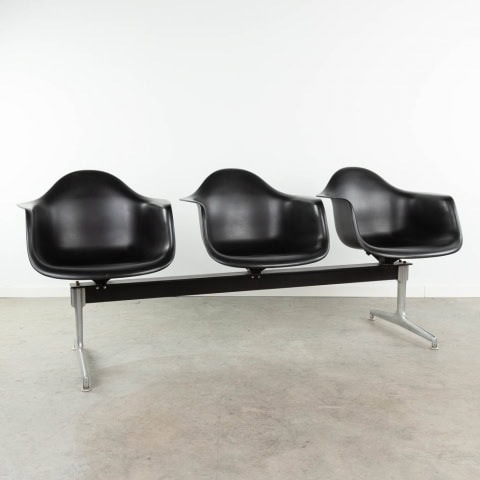 Drie moderne, zwarte, gegoten kunststof stoelen met armleuningen zijn op een rij met elkaar verbonden door een metalen balk met zilverkleurige poten. Ze staan tegen een eenvoudige witte muur en een betonnen vloer.