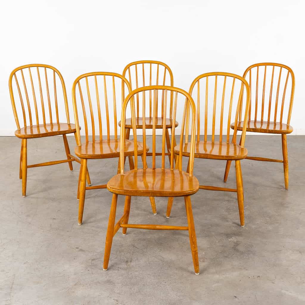 Zes houten eetkamerstoelen met gebogen, spindelvormige rugleuningen en ronde zittingen, gerangschikt in twee rijen op een betonnen vloer met een effen witte achtergrond.