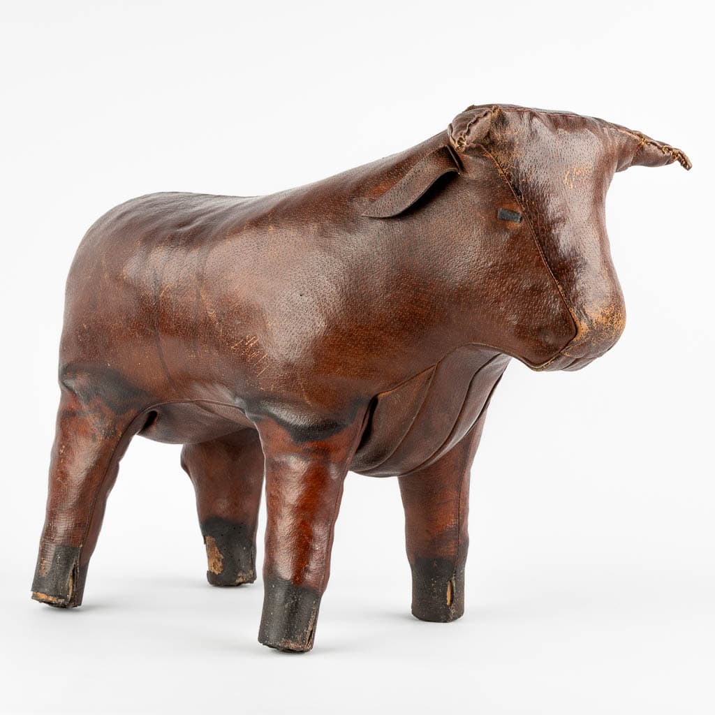 Een vintage leren voetenbankje in de vorm van een stier, met een versleten bruin oppervlak en eenvoudige, ronde kenmerken. De poef staat op vier stevige poten tegen een witte achtergrond.