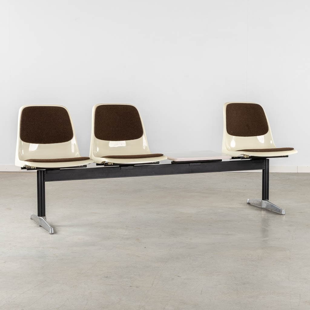 Drie aan elkaar gekoppelde plastic stoelen met bruine kussens en metalen poten staan op een rij, met een lege ruimte tussen de middelste en de rechterstoel, op een kale vloer tegen een eenvoudige witte muur.