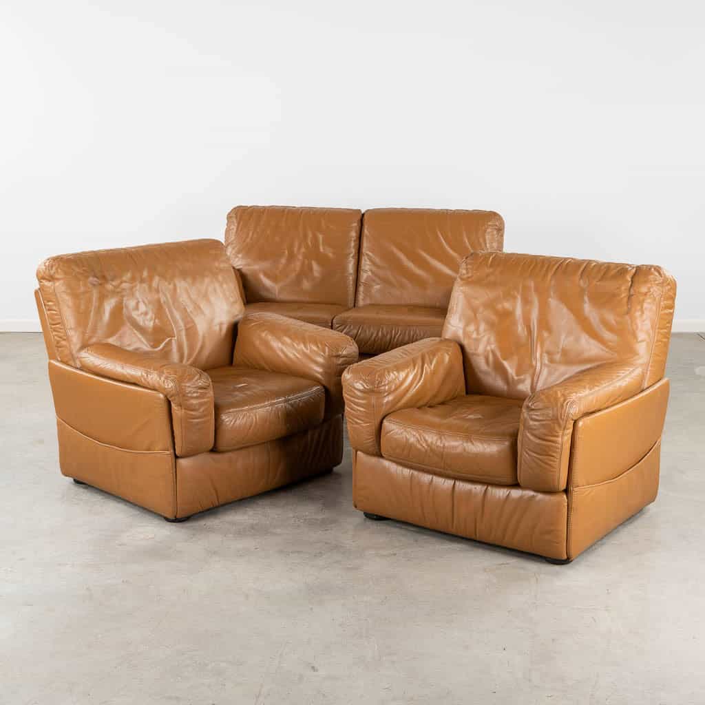 Een set van drie vintage bruine leren fauteuils naast elkaar op een lichtgrijze betonnen vloer tegen een effen witte muur. De stoelen hebben een licht versleten, luxueuze uitstraling.