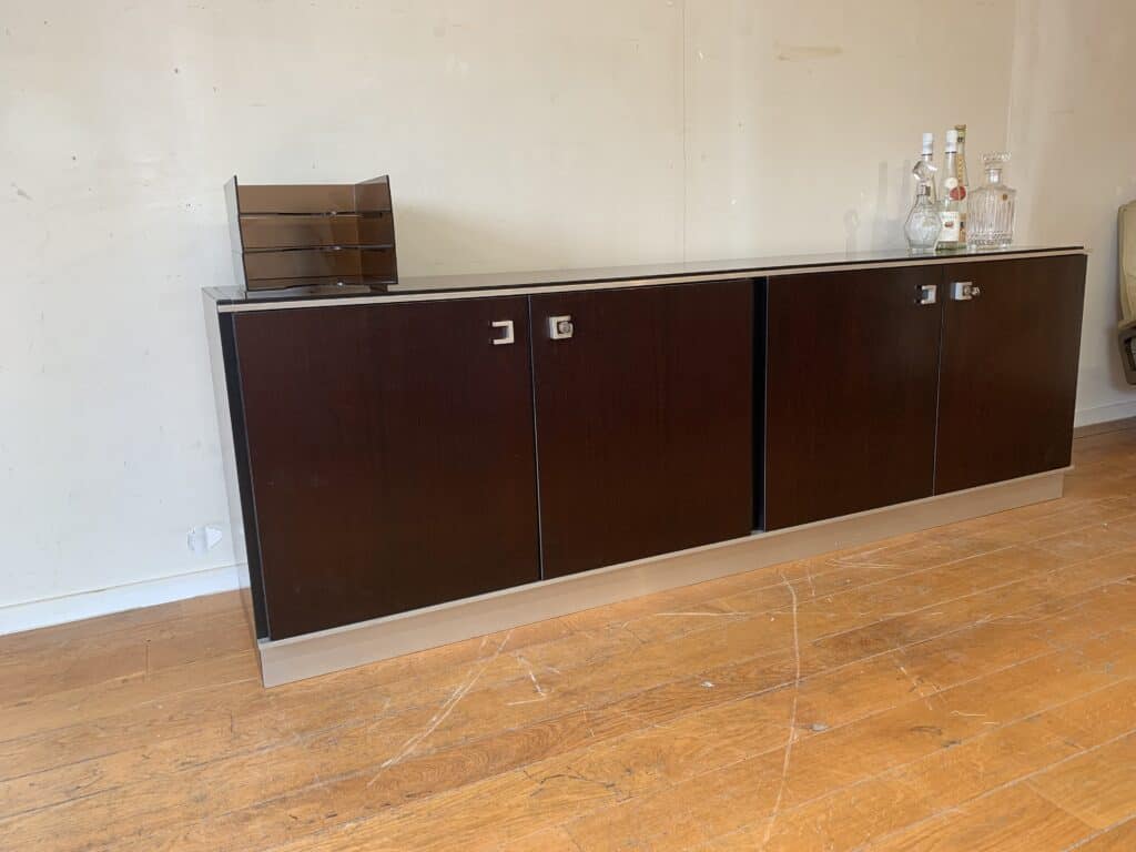 Een donkerhouten dressoir staat tegen een beige muur op een houten vloer. Bovenop het dressoir staan verschillende glazen flessen en een kleine donkere ordner.