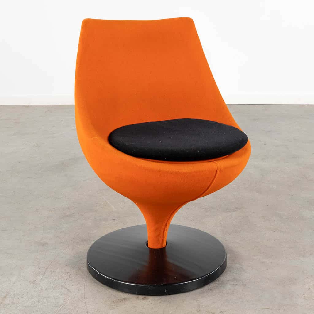 Een moderne, oranje draaistoel met een gebogen rugleuning, een zwart rond zitkussen en een ronde, zwarte voet, geplaatst op een betonnen vloer tegen een effen witte muur.