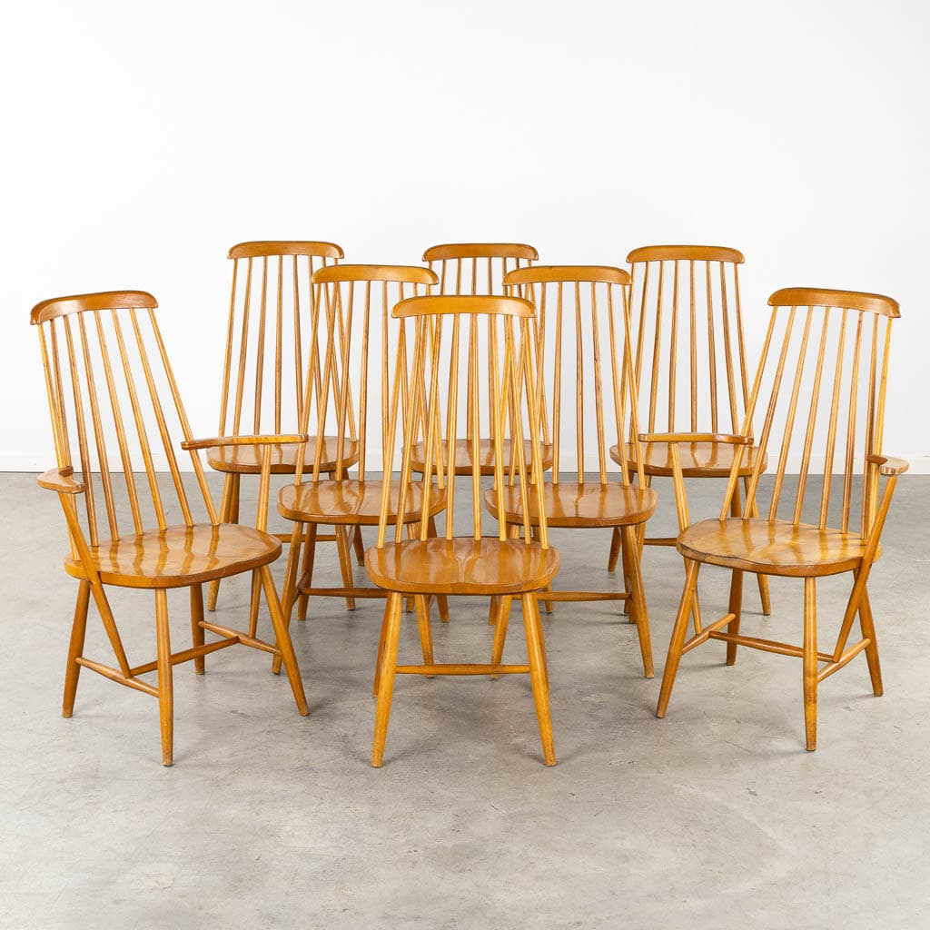 Acht houten stoelen met spijlenrug staan in een halve cirkel op een betonnen vloer, met een effen witte muur op de achtergrond. De stoelen hebben hoge, slanke rugleuningen en eenvoudige, taps toelopende poten.