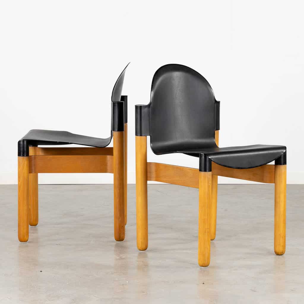 Twee moderne stoelen met gebogen, zwarte zittingen en rugleuningen, en lichte houten poten en frames, staan naast elkaar op een lichtgrijze vloer tegen een eenvoudige witte muur.
