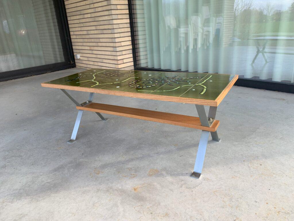 Een moderne rechthoekige tafel met een groen blad met een landkaartmotief, een houten plankje eronder en schuine metalen poten, geplaatst op een betonnen terras bij grote glazen deuren.