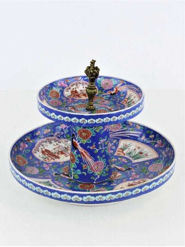 Een tweedelig porseleinen serveerblad met ingewikkelde blauwe, rode en witte bloemen- en landschapspatronen, met een decoratief messing handvat in het midden.