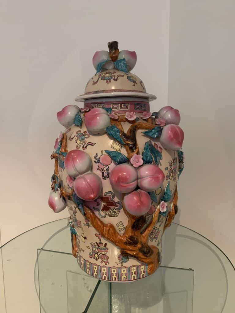 Een decoratieve porseleinen pot met deksel, versierd met reliëf, kleurrijke perziken, takken en bloemen. De pot is versierd met ingewikkelde patronen en motieven in roze, blauw en bruintinten, weergegeven op een glazen oppervlak.
