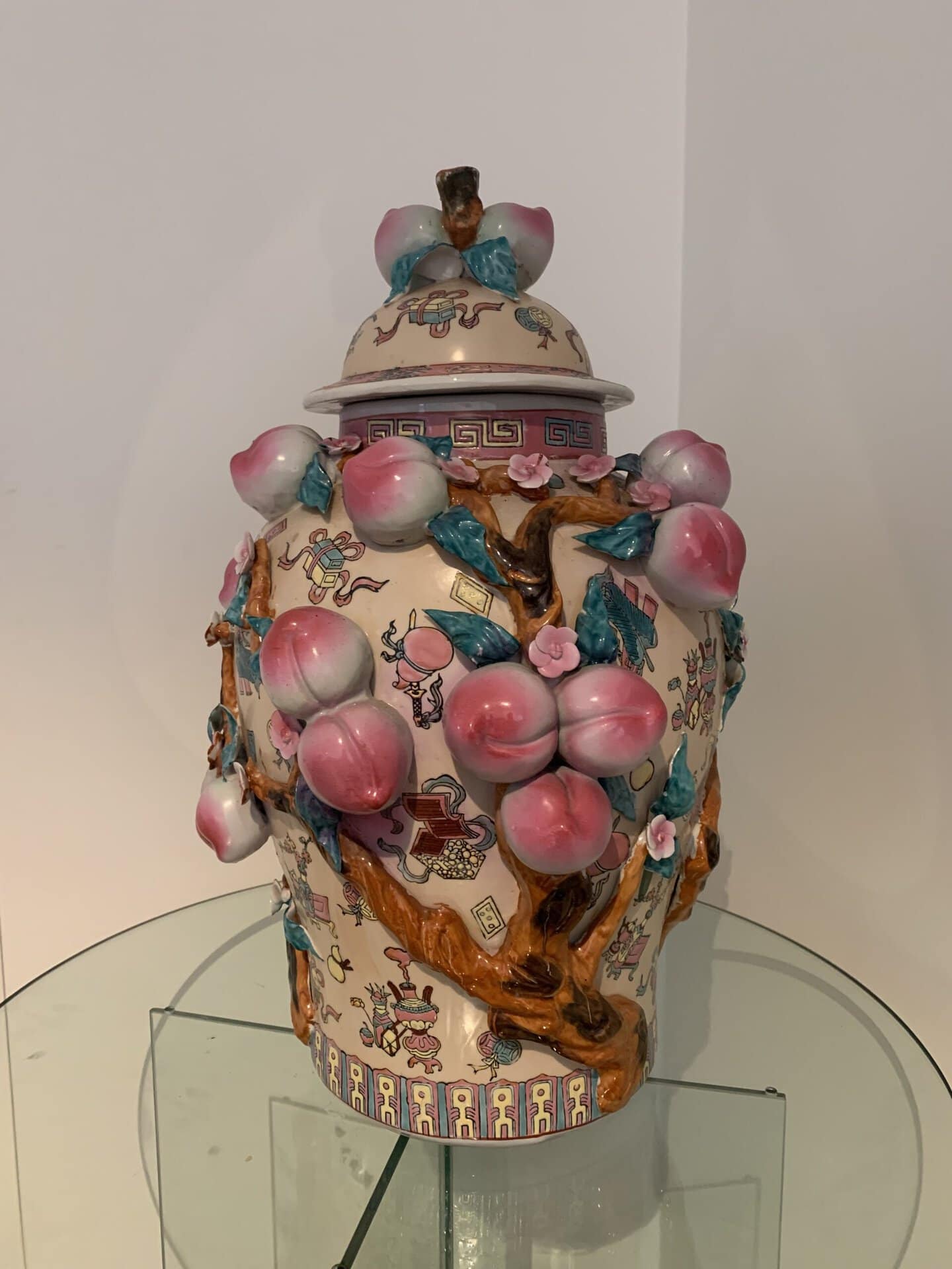 Een decoratieve porseleinen pot met deksel, versierd met reliëf, kleurrijke perziken, takken en bloemen. De pot is versierd met ingewikkelde patronen en motieven in roze, blauw en bruintinten, weergegeven op een glazen oppervlak.