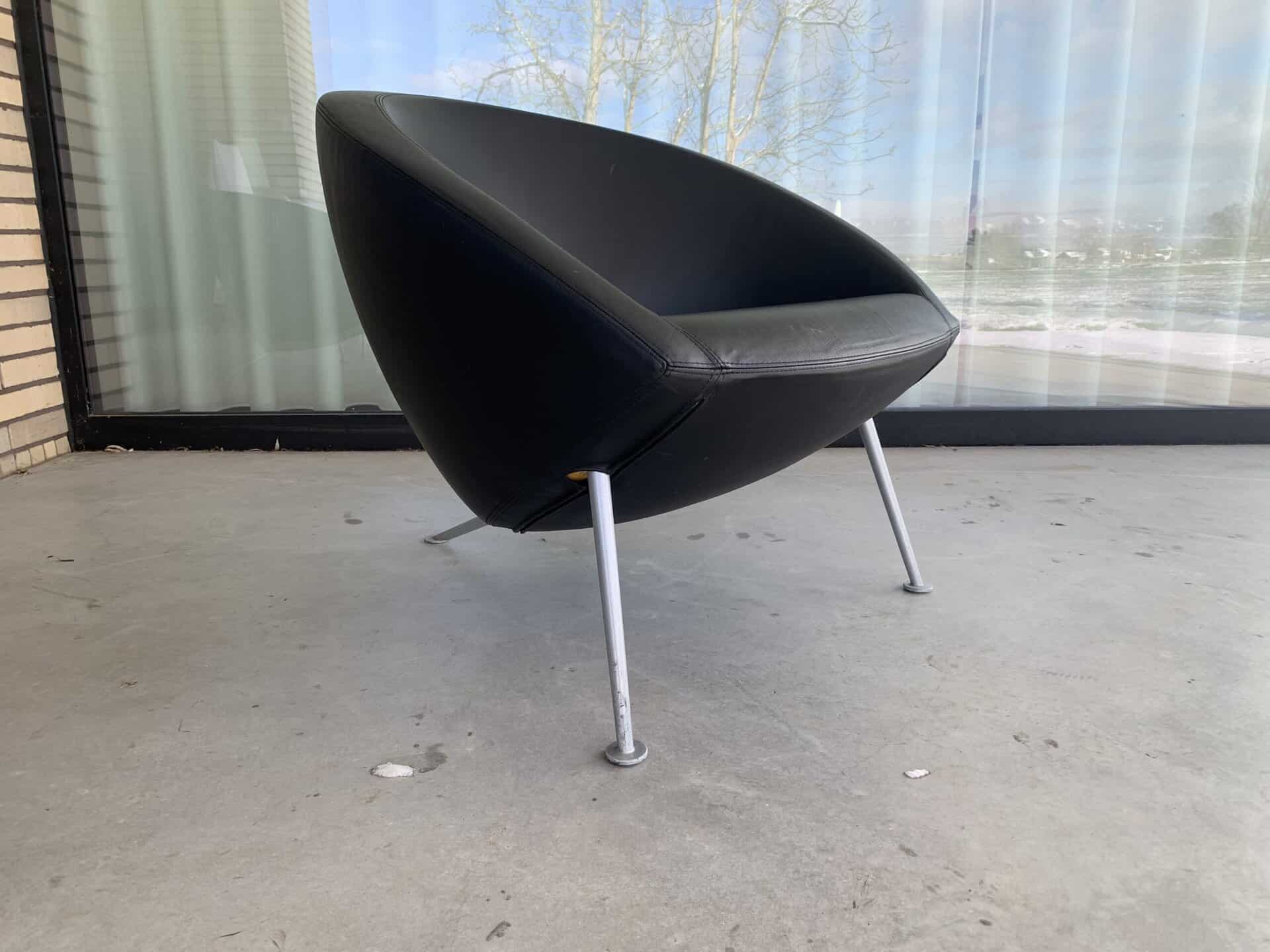 Een moderne, zwarte driehoekige stoel met drie metalen poten staat op een betonnen vloer voor een groot raam met witte gordijnen en uitzicht op de bomen buiten.