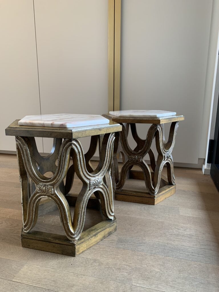 Twee sierlijke zeshoekige bijzettafeltjes met goudkleurige, gesneden houten onderstellen en witte marmeren bladen, geplaatst op een lichte houten vloer met een witte kast en spiegel op de achtergrond.