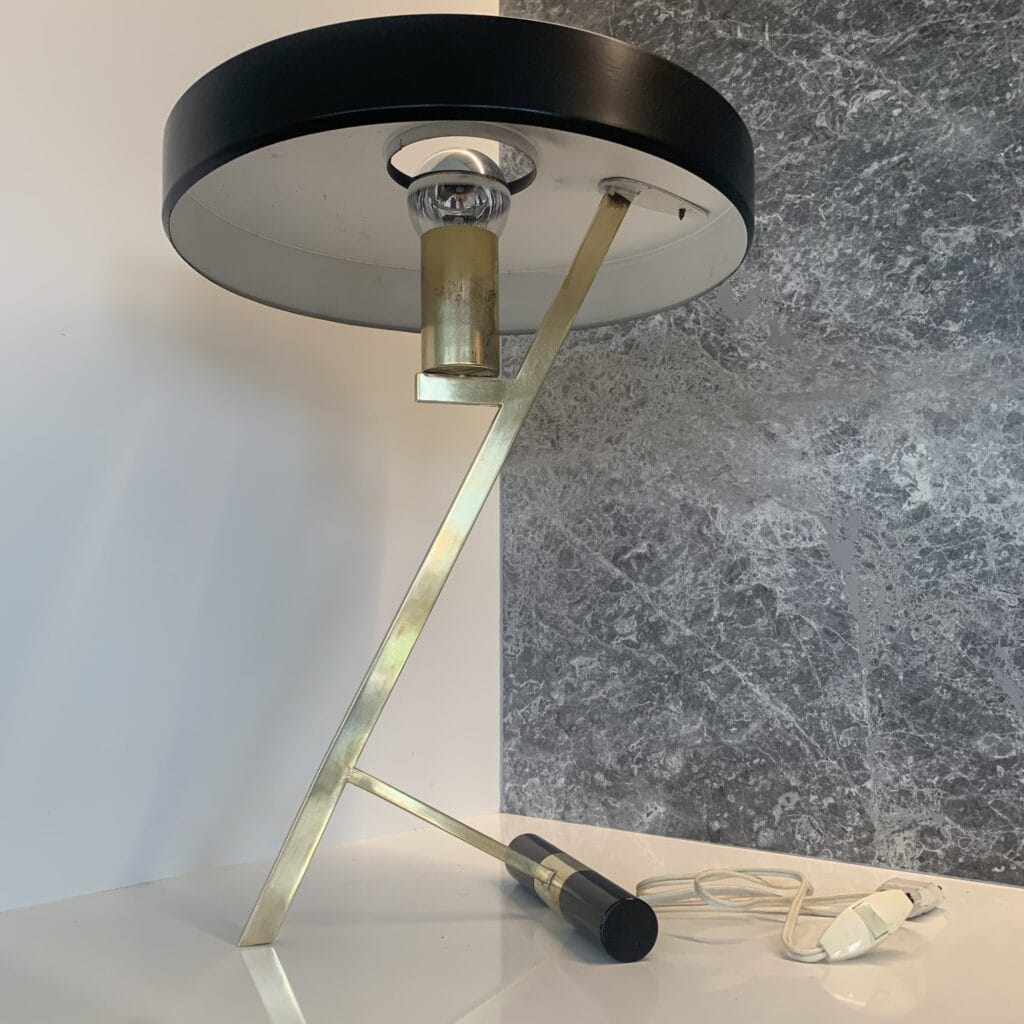 Een moderne tafellamp met een ronde zwarte kap, messing hoekige poten en een zichtbare gloeilamp, geplaatst op een wit oppervlak tegen een grijs gemarmerde muur. Het netsnoer en de stekker zijn zichtbaar op de tafel.