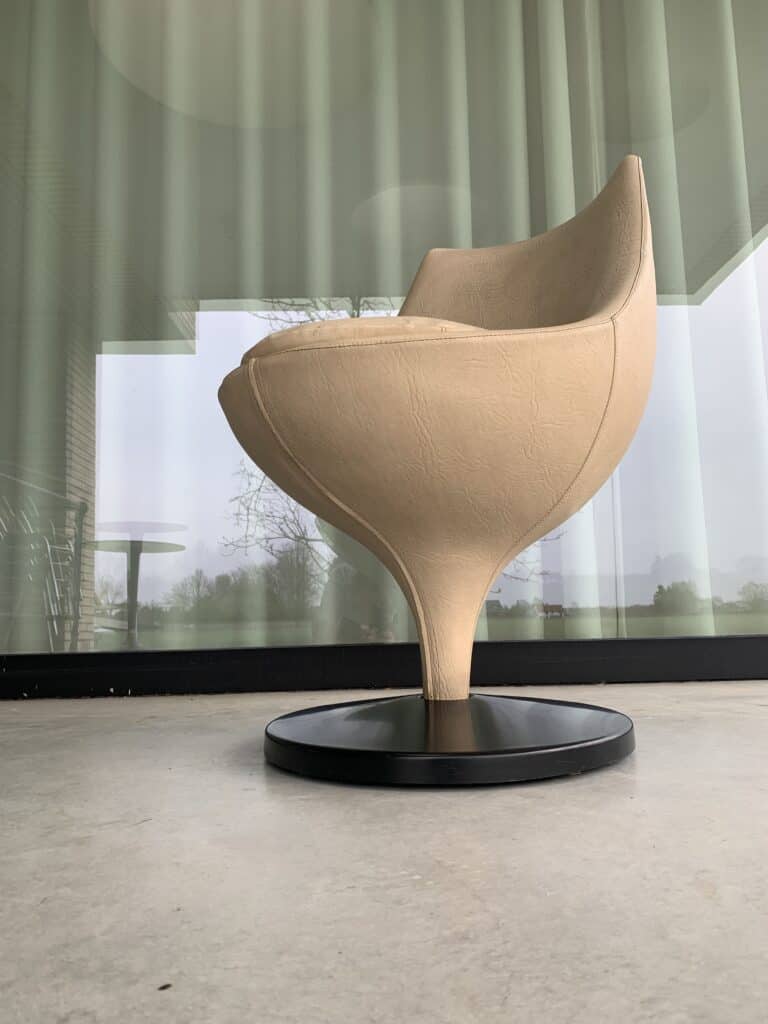 Een moderne, beige, tulpvormige stoel met een ronde basis staat op een gladde betonnen vloer voor hoge glazen ramen met groene gordijnen.