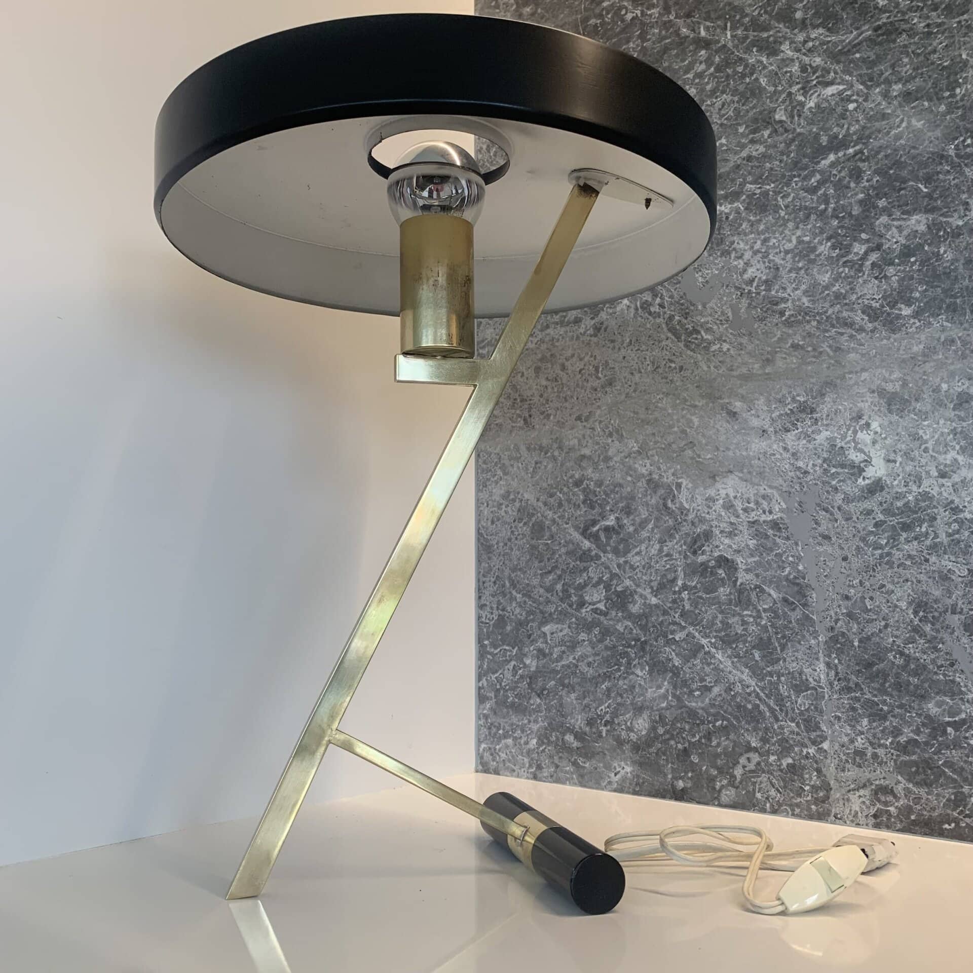 Een moderne tafellamp met een ronde zwarte kap, messing hoekige poten en een zichtbare gloeilamp, geplaatst op een wit oppervlak tegen een grijs gemarmerde muur. Het netsnoer en de stekker zijn zichtbaar op de tafel.