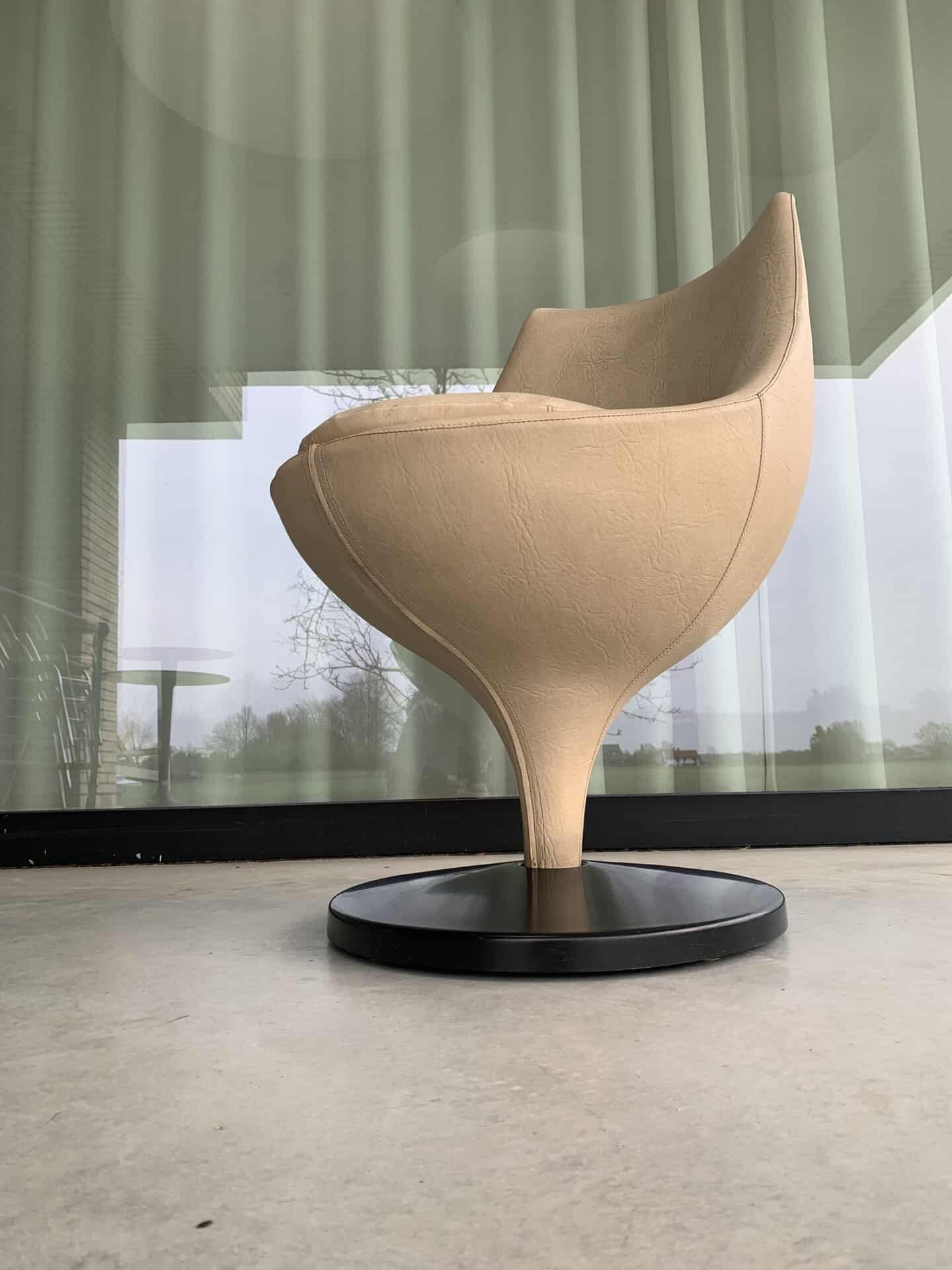 Een moderne, beige, tulpvormige stoel met een ronde basis staat op een gladde betonnen vloer voor hoge glazen ramen met groene gordijnen.