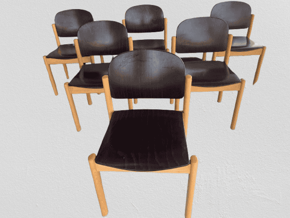 Zes houten stoelen met donkere, gebogen rugleuningen en zittingen staan in twee rijen op een witte achtergrond. De voorste stoel staat iets los van de andere.