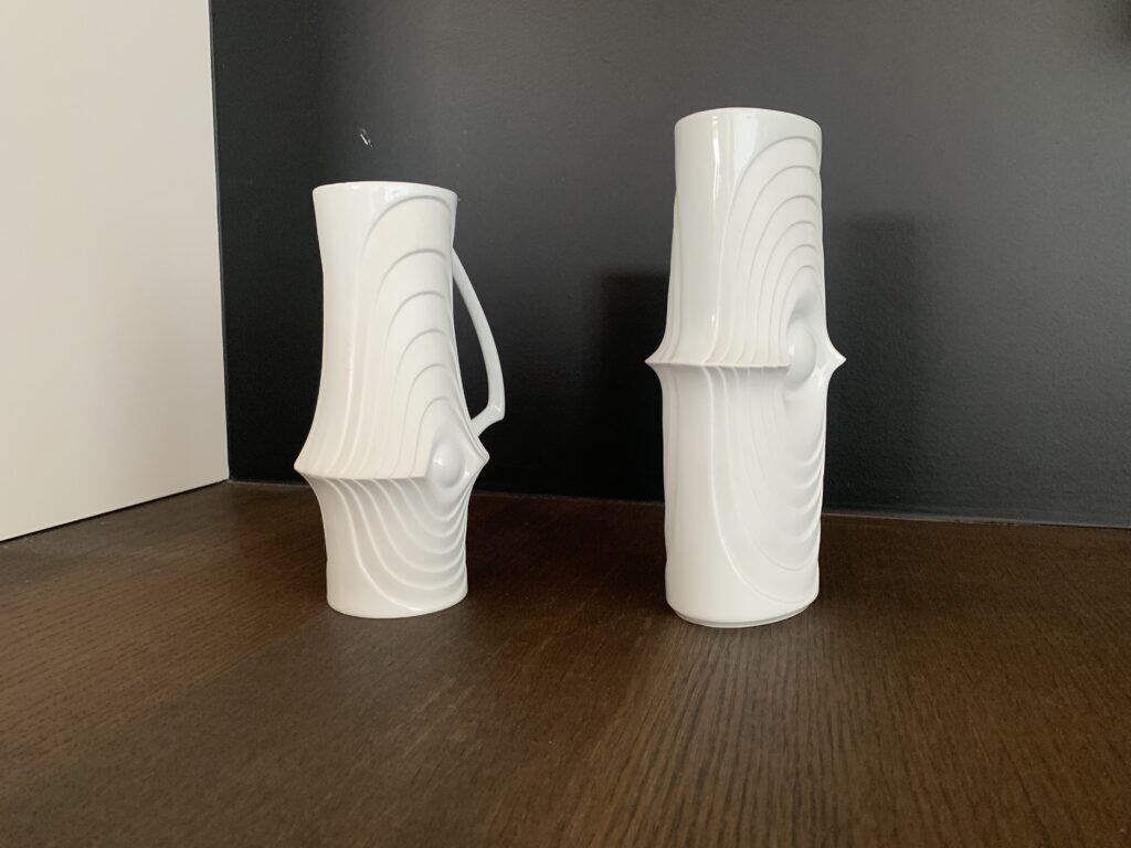 Twee witte keramische vazen met golvende, concentrische patronen en uitstulpingen staan op een donker houten oppervlak tegen een zwarte achtergrond. De ene vaas heeft een handvat, de andere is hoger en cilindrisch.