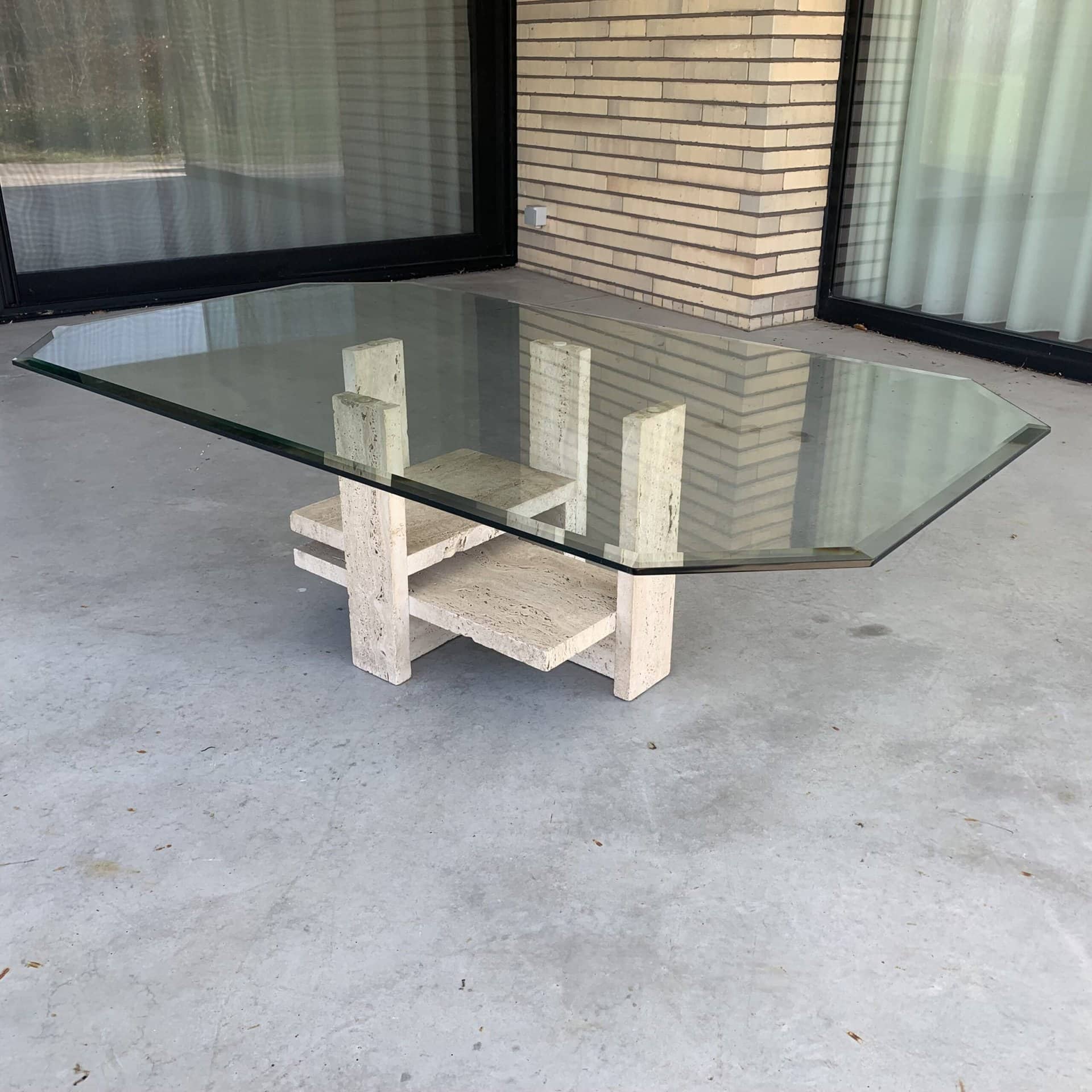 Een moderne salontafel met een dik, rechthoekig glazen blad en een geometrisch stenen onderstel, geplaatst op een betonnen vloer voor ramen met witte gordijnen en een beige bakstenen muur.