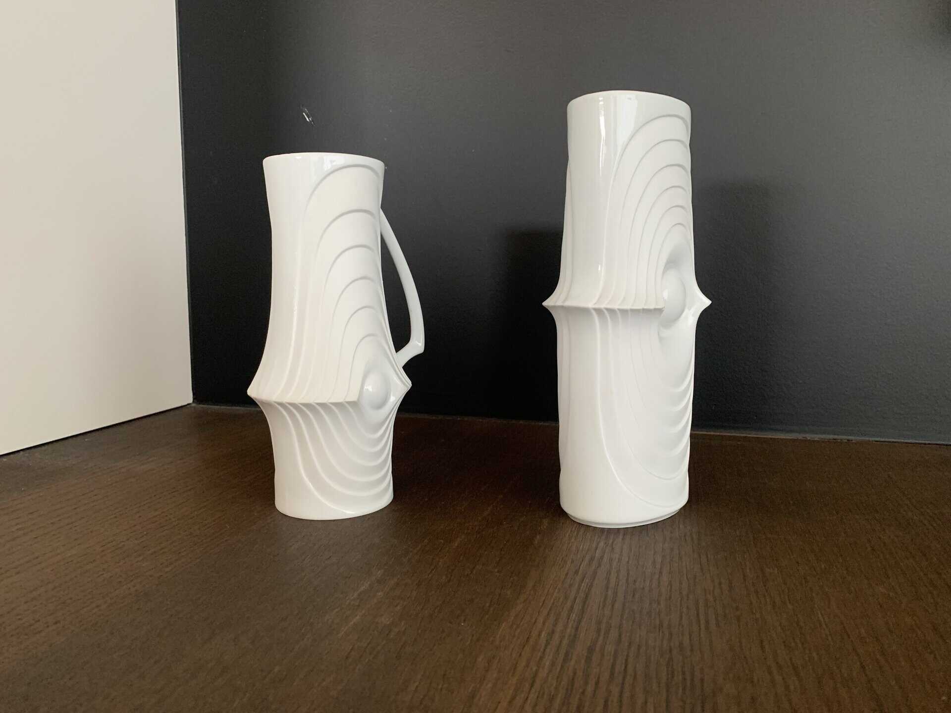 Twee witte keramische vazen met golvende, concentrische patronen en uitstulpingen staan op een donker houten oppervlak tegen een zwarte achtergrond. De ene vaas heeft een handvat, de andere is hoger en cilindrisch.