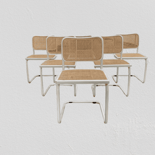 Vier moderne stoelen met een wit metalen frame en een zitting en rugleuning van gevlochten rotan staan in twee rijen tegen een eenvoudige witte muur.
