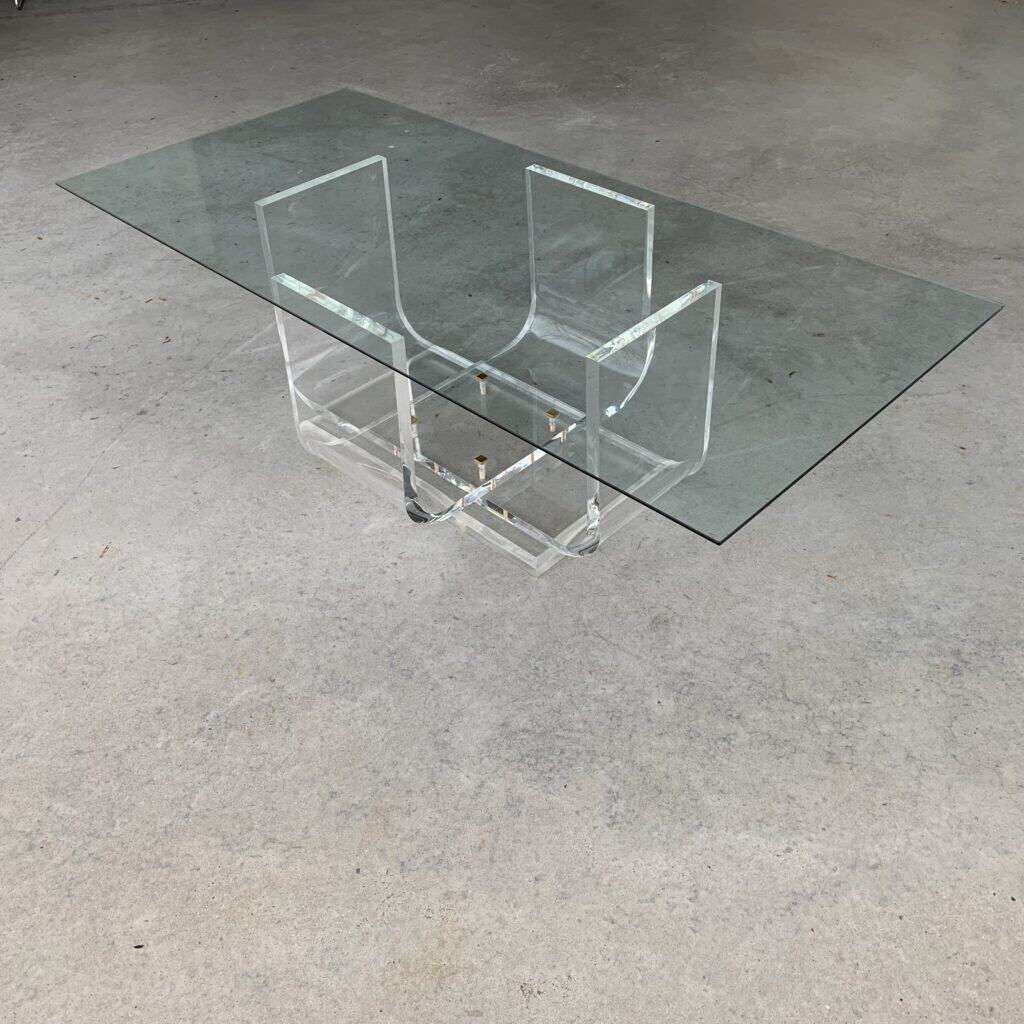 Een moderne rechthoekige glazen salontafel met een transparant onderstel van acryl met geometrische vormen, geplaatst op een gladde betonnen vloer.