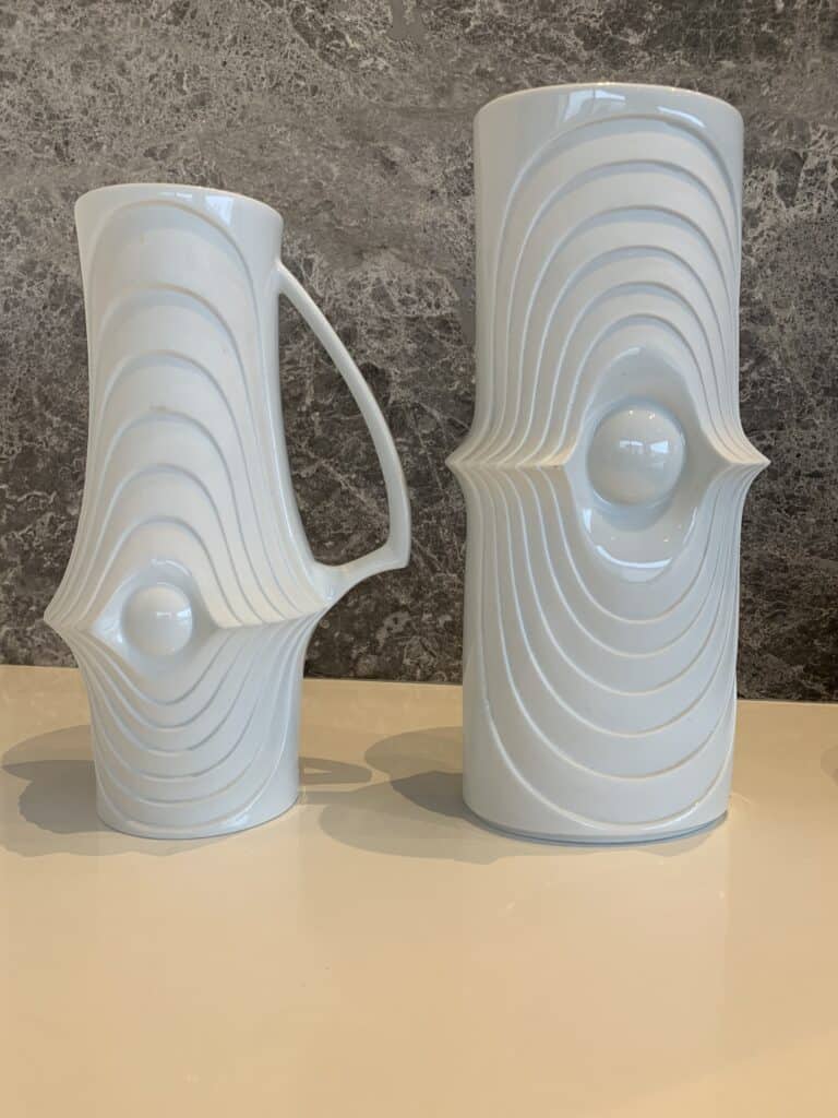 Twee witte keramische vazen met gebeeldhouwde, symmetrische ribbels en een centraal bolvormig ontwerp, tentoongesteld op een licht oppervlak tegen een getextureerde grijze stenen achtergrond. De ene vaas heeft een handvat; de andere is hoger en cilindrisch.