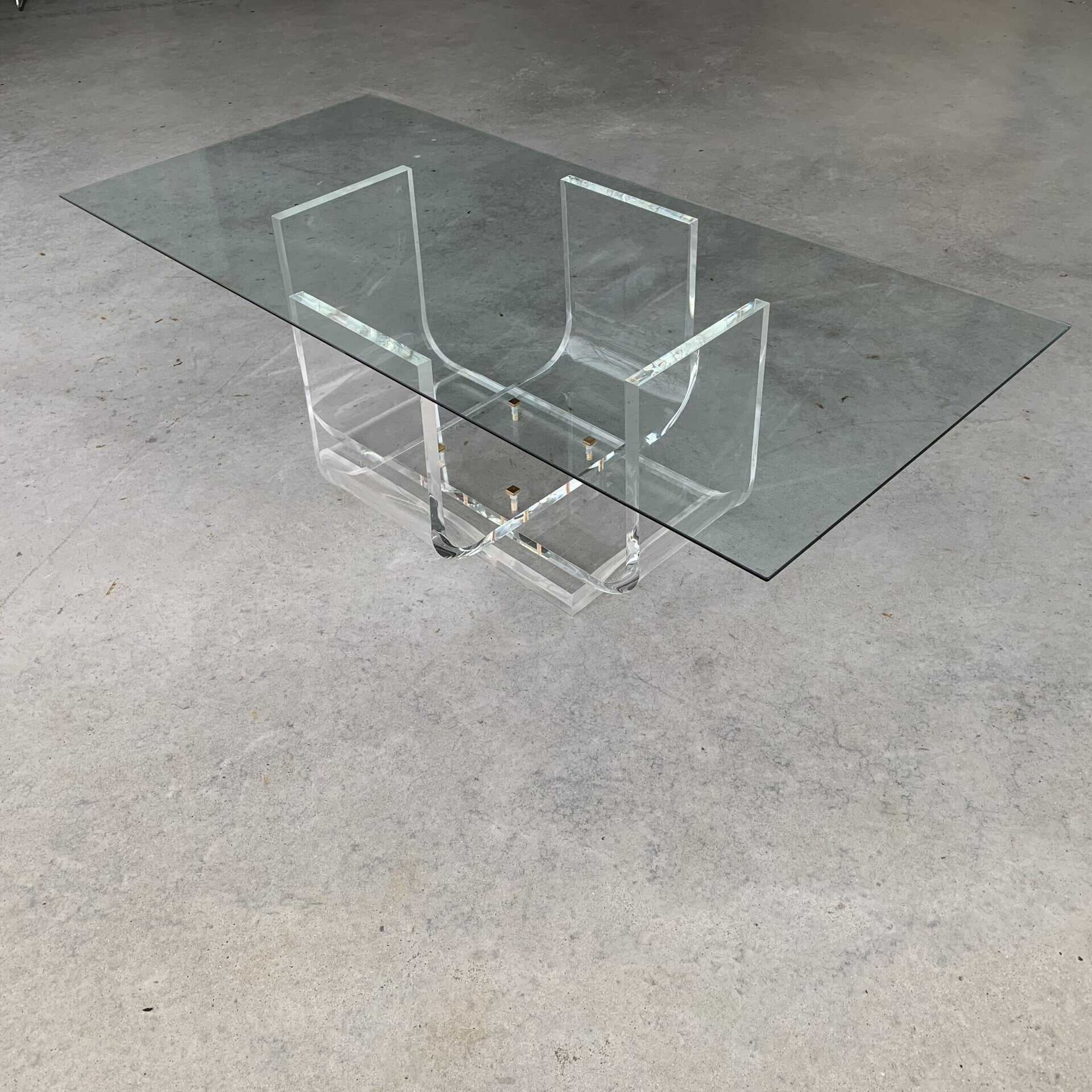 Een moderne rechthoekige glazen salontafel met een transparant onderstel van acryl met geometrische vormen, geplaatst op een gladde betonnen vloer.