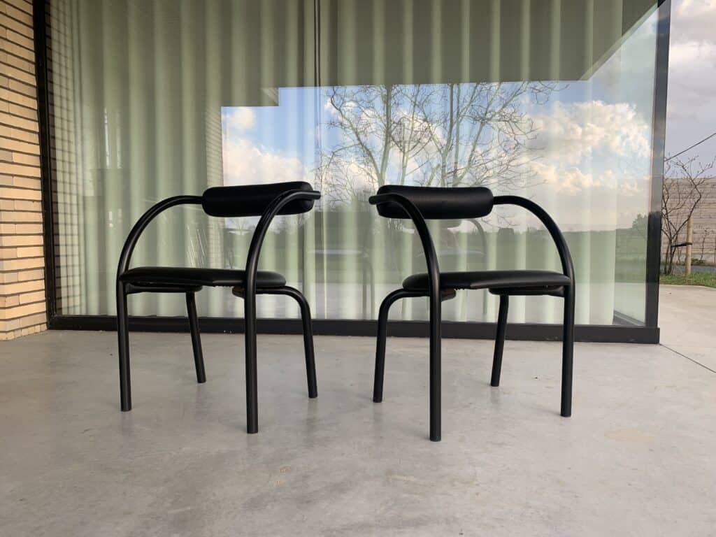 Twee moderne zwarte stoelen met gebogen rugleuningen en zittingen staan op een betonnen terras voor een groot raam met weerspiegelingen van bomen en lucht. De achtergrond bestaat uit verticale jaloezieën en een gedeeltelijk bakstenen muur.