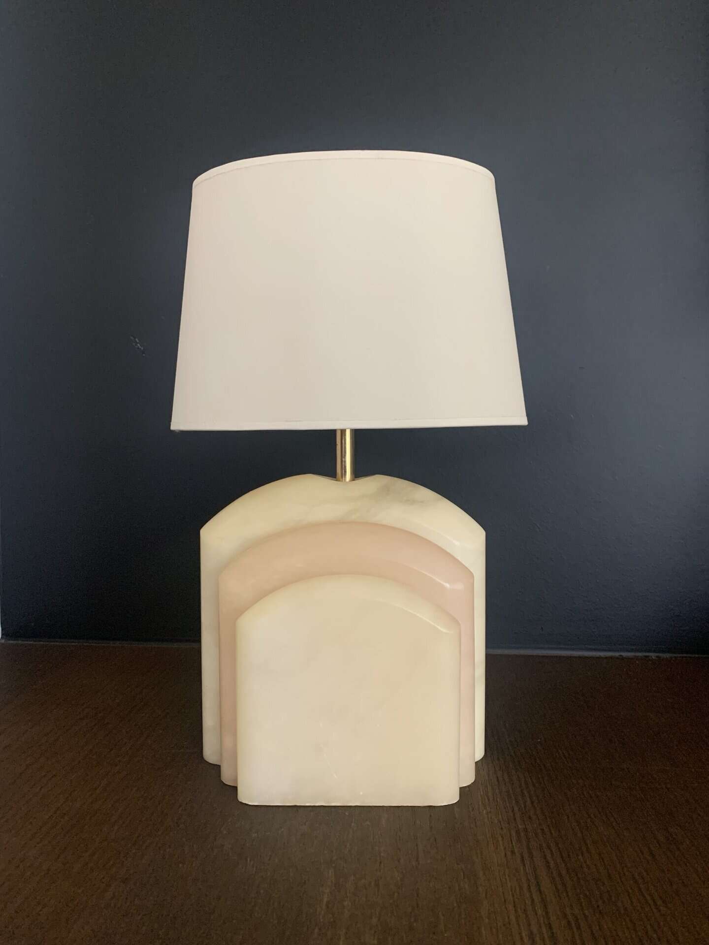 Een tafellamp met een witte lampenkap en een marmeren voet in art-decostijl met gelaagde, boogvormige delen, tegen een donkere muur op een houten oppervlak.