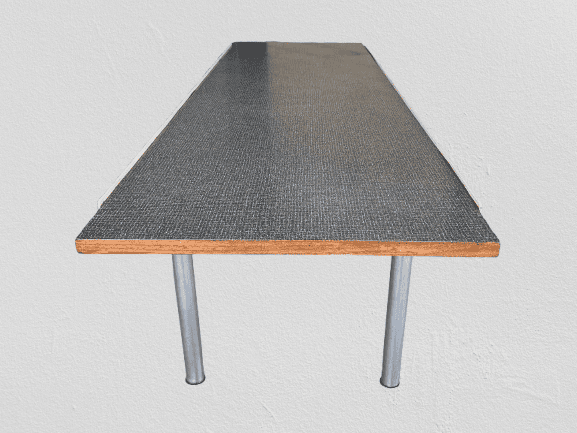 Een lange rechthoekige tafel met een donker oppervlak, houten randen en twee zichtbare metalen poten, tegen een eenvoudige lichtgekleurde muur.