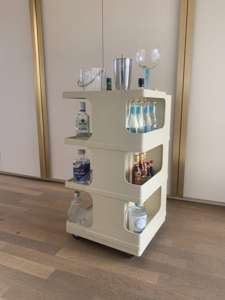 Een beige rolwagen met drie planken biedt plaats aan flessen alcohol, mixers, glaswerk en cocktailaccessoires. De wagen staat op een houten vloer in een moderne ruimte met gouden en witte accenten.