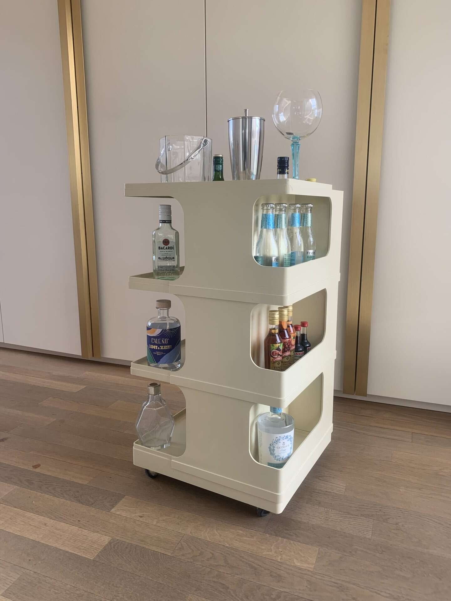Een beige rolwagen met drie planken biedt plaats aan flessen alcohol, mixers, glaswerk en cocktailaccessoires. De wagen staat op een houten vloer in een moderne ruimte met gouden en witte accenten.