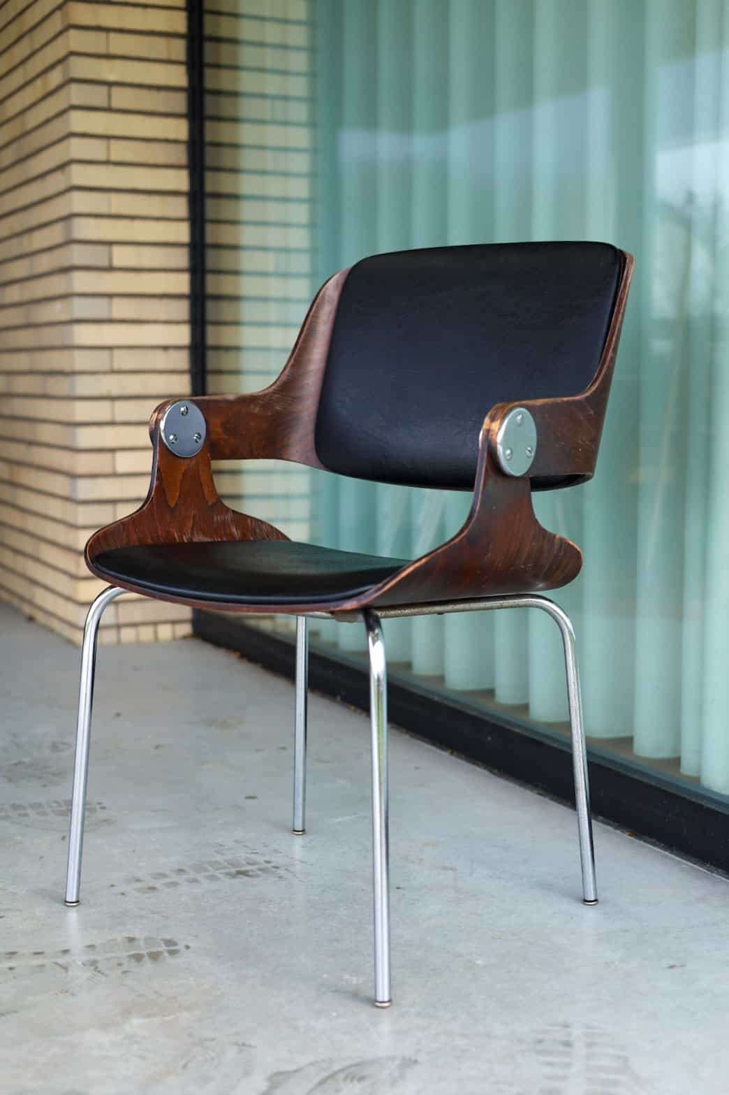 Een moderne stoel met een donker houten frame, zwarte zitting en rugleuning en metalen poten staat op een betonnen vloer bij een glazen raam met verticale gordijnen. Op de achtergrond is een bakstenen muur zichtbaar.