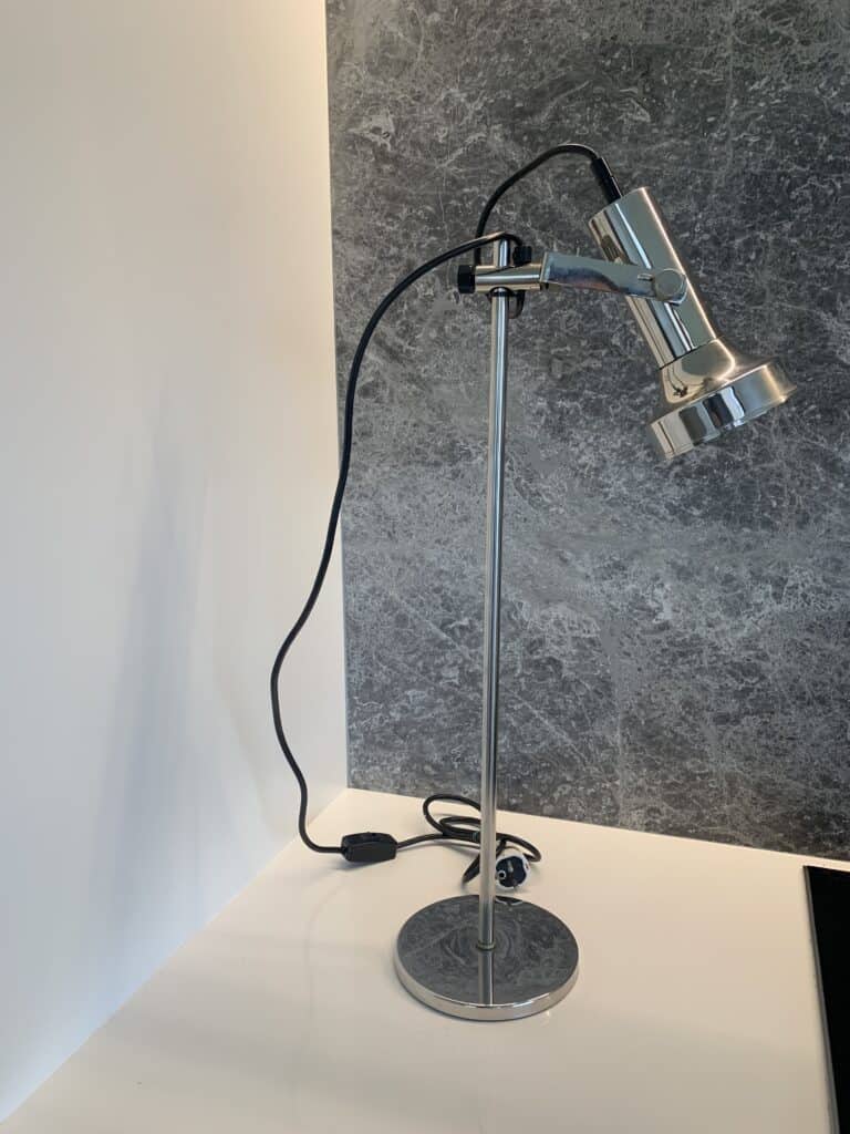 Een zilveren, verstelbare bureaulamp met een zwart snoer staat op een wit oppervlak tegen een grijs gemarmerde muur. De lamp heeft een moderne stijl en is aangesloten op het lichtnet, met het snoer losjes opgerold aan de voet.