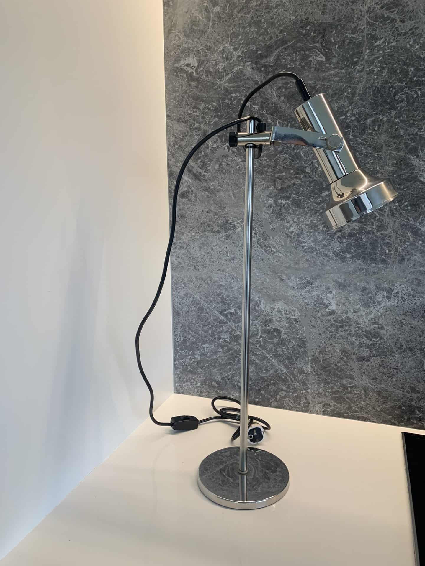 Een zilveren, verstelbare bureaulamp met een zwart snoer staat op een wit oppervlak tegen een grijs gemarmerde muur. De lamp heeft een moderne stijl en is aangesloten op het lichtnet, met het snoer losjes opgerold aan de voet.